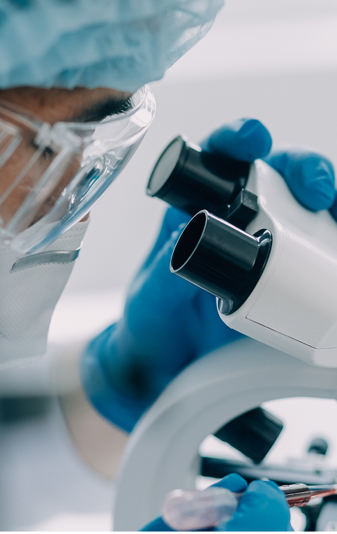 Close-up of a laboratory technician wearing protective eyewear and gloves, examining a sample through a microscope in a clinical setting.